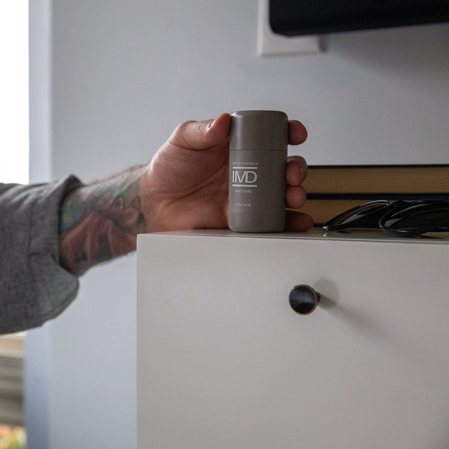 Tattooed person reaching for IMD Daily Care Tattoo Balm on a bedroom dresser — daily aftercare to keep colored ink vibrant