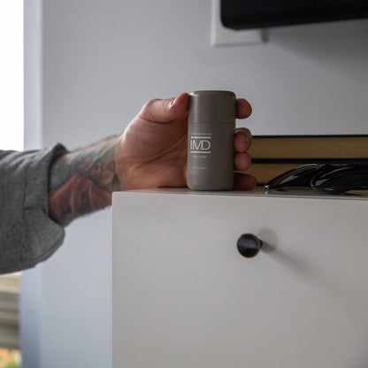 Tattooed person reaching for IMD Daily Care Tattoo Balm on a bedroom dresser — daily aftercare to keep colored ink vibrant